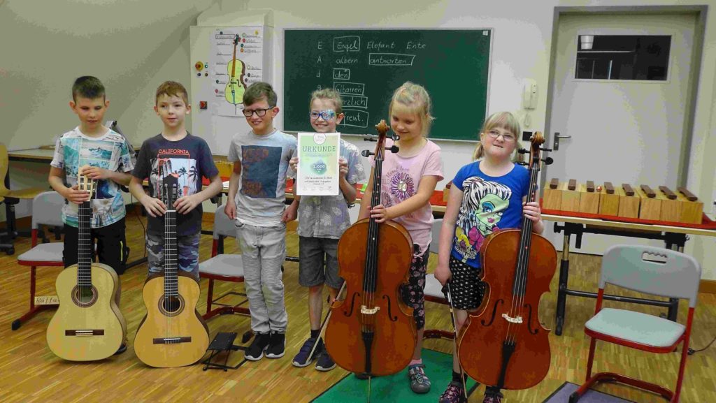Sechs Grundschüler haben sich mit zwei Gitarren und zwei Celli im Probenraum Haus 16 aufgestellt und freuen sich über die Urkunde zum Preis.)