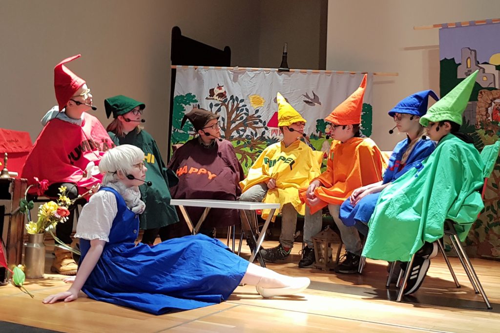 Schauspiel-Nachwuchs auf der Bühne: Sieben Zwerge in knallbunten Kostümen mit Zipfelmützen und im Vordergrund eine Prinzessin. Alle tragen ein Headset-Mikrofon.