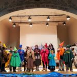 Diese Bild zeigt die Theatergruppe auf der Bühne. Darüber ein Schriftzug: "Frisch, Fromm, Fröhlich, Frei, sei die ganze Turnerei."