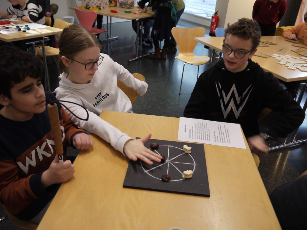 Drei Schüler sitzen um einen Tisch und Spielen das Spiel „Kreismühle“. Als Spielfiguren werden Kastanien verwendet.