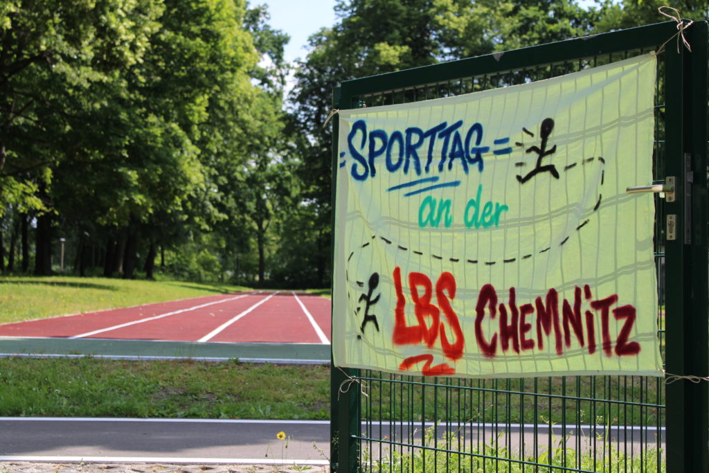 Ein handgemalter Banner zeigt die Aufschrift "Sporttag der LBS Chemnitz". Dahinter ist der Sportplatz zu sehen.