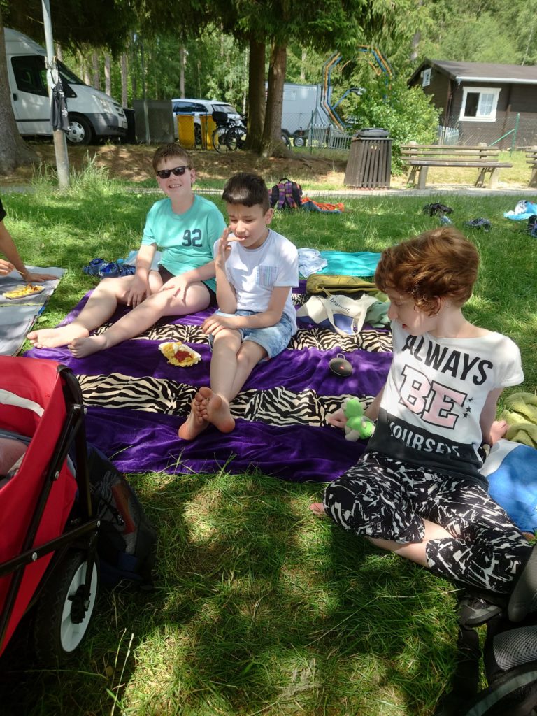 Eine entspannte Szene im Grünen: drei Kinder genießen das gute Wetter auf der Wiese. Sie sitzen auf Decken und Badetüchern. Im Hintergrund stehen Camp-Mobile und Bungalows.