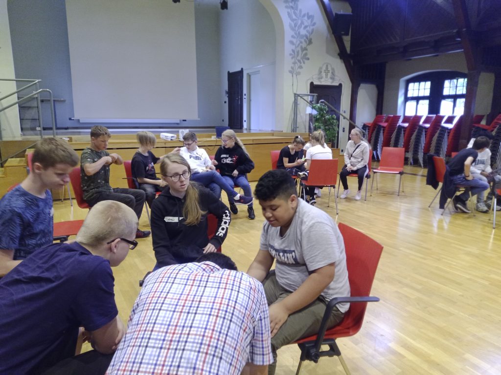 Eine Aufnahme des gesamten Raumes zeigt ein Dutzend Schüler, die in Gruppen sitzen und miteinander sprechen. Alle wirken vertieft und unterhalten sich angeregt.