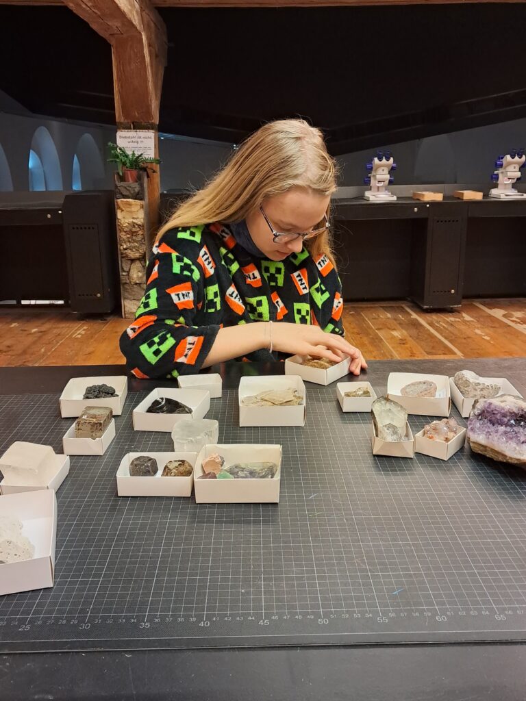 Eine Schülerin sitzt am Tisch und untersucht kleine Kästchen mit verschiedenen Materialien