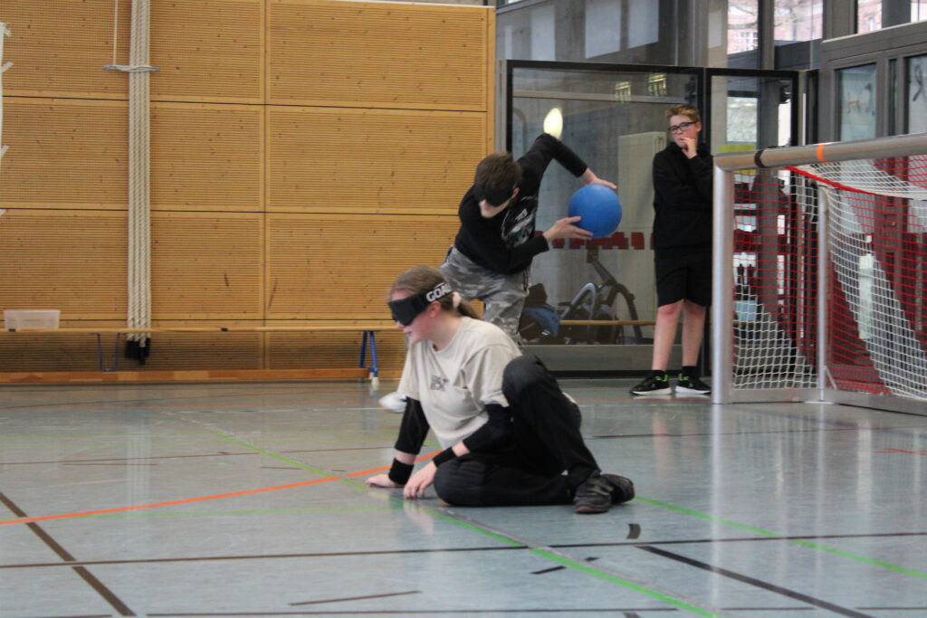Ein Schüler wirft den Goalball und seine Mitspielerin befindet sich in der Verteidigungsposition. Beide tragen Dunkelbrillen.