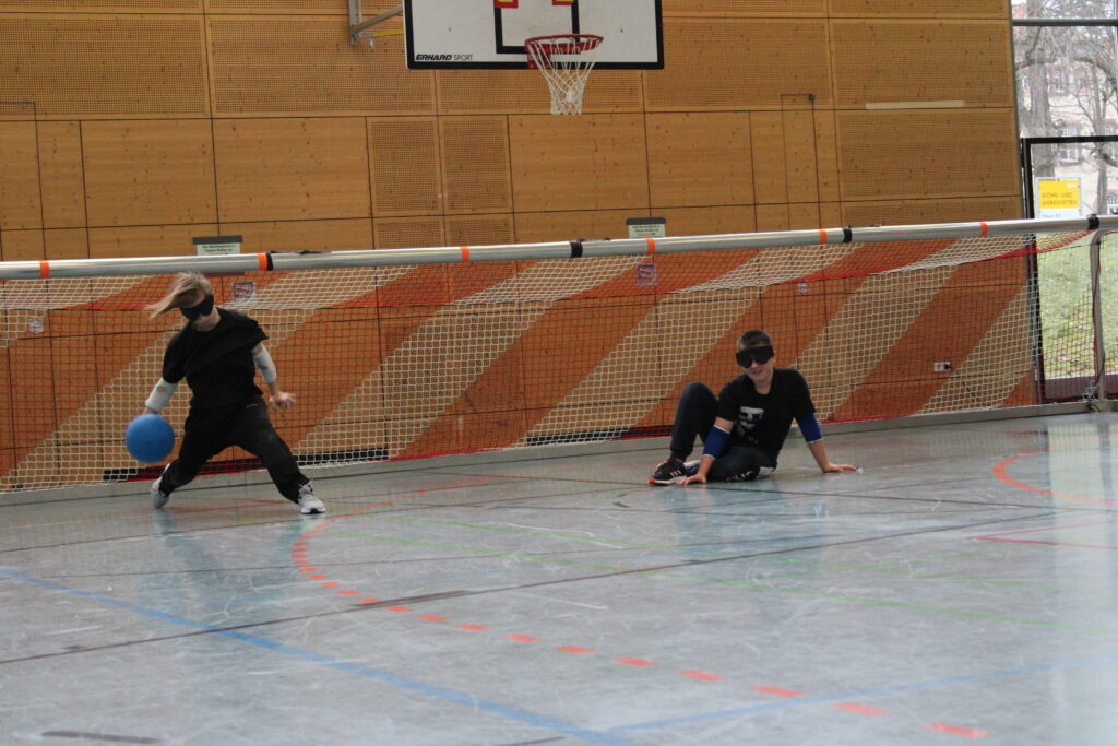 Ein Schüler wirft den Goalball und sein Mitspieler befindet sich in der Verteidigungsposition. Beide tragen Dunkelbrillen.