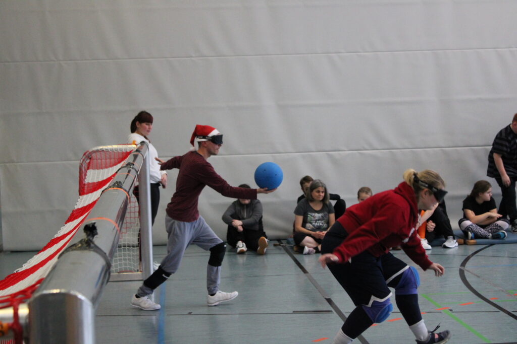 Ein Lehrer hält den Goalball in seiner Hand und ist bereit für den Angriffswurf. Seine Teamkollegin geht gerade in die Verteidigungsposition.
