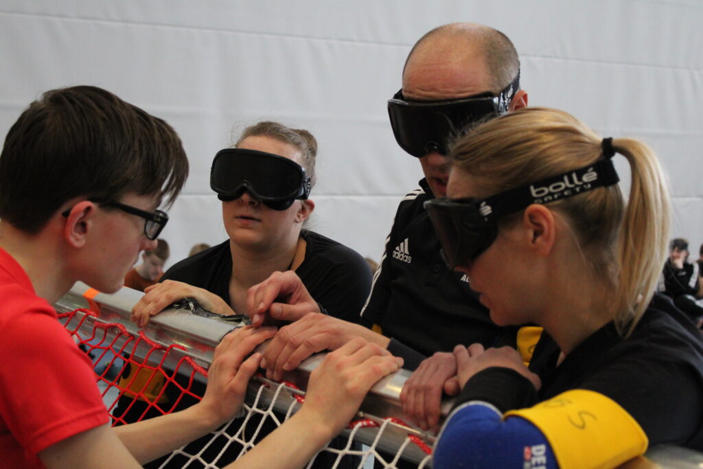 Während einer Spielpause versammelt sich ein Lehrerteam am Torpfosten und bespricht sich mit ihrem Trainer. Die Spieler tragen Dunkelbrillen.