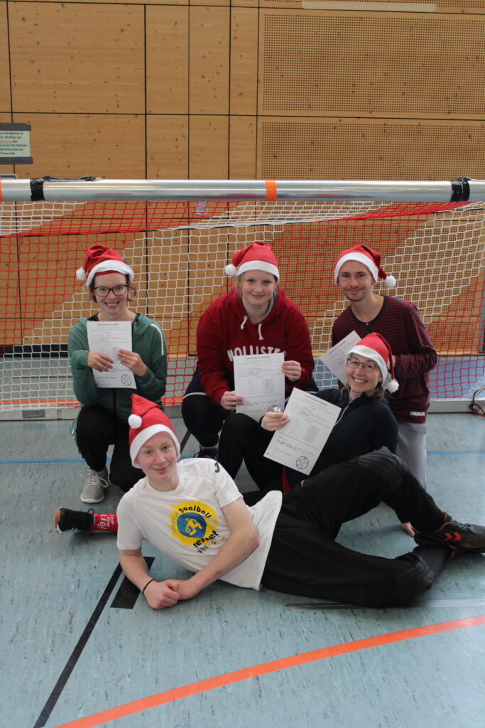 Eines der Lehrerteams posiert vor dem Goalballtor mit ihrem Trainer (ein Schüler). Alle tragen Weihnachtsmützen auf dem Kopf.