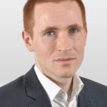 Stefan sitzt mit weißem Hemd, schwarzem Sakko und kurzen roten Haaren vor der Kamera.