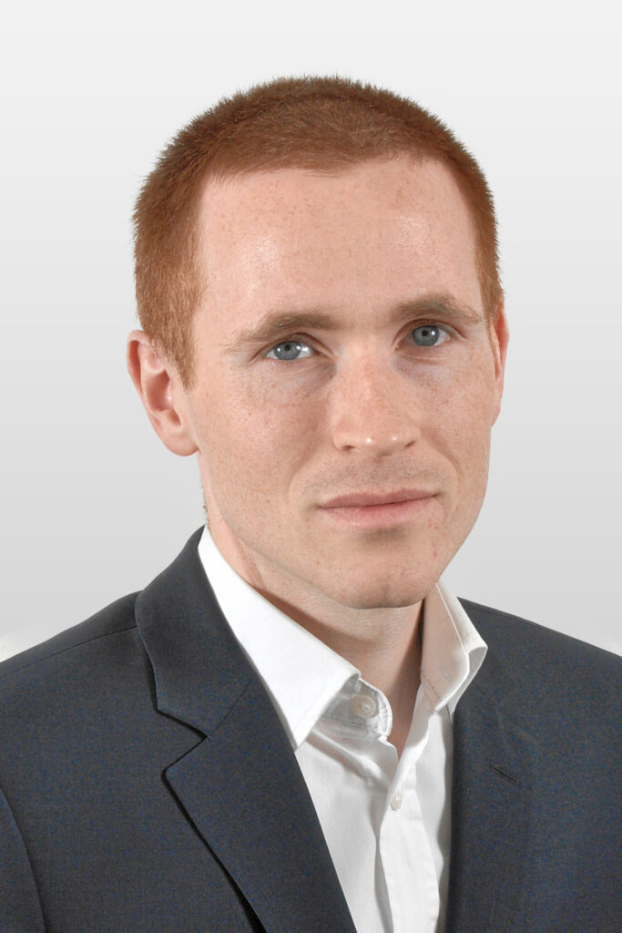 Stefan sitzt mit weißem Hemd, schwarzem Sakko und kurzen roten Haaren vor der Kamera.