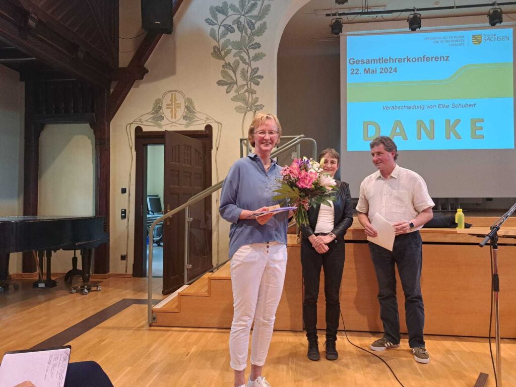 Frau Schubert lächelt und hält einen Umschlag und einen Strauß Blumen in den Händen