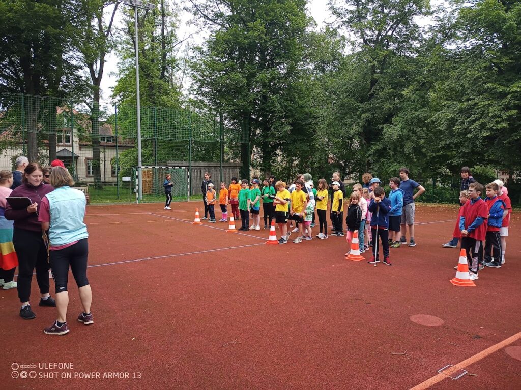 Die Schülerinnen und Schüler stehen auf dem Sportplatz. Sie sind nebeneinander in Reihen aufgestellt und hören gespannt zu.
