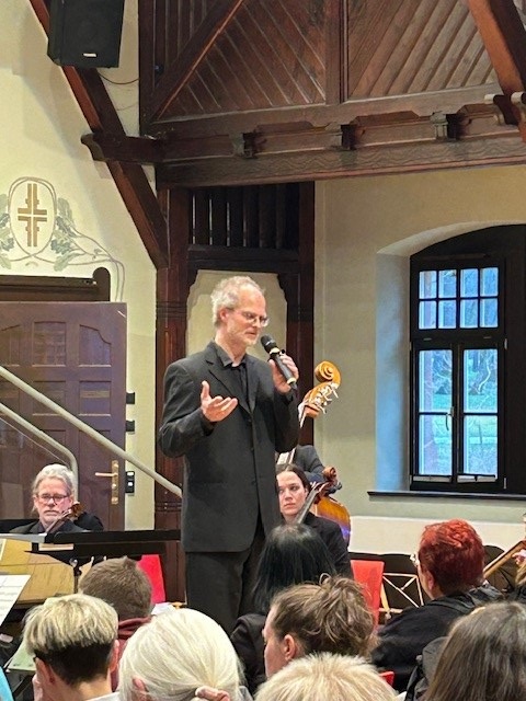 Ein Sänger mit Mikrofon steht inmitten der sitzenden Musizierenden und singt.