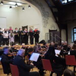 Im Vordergrund sitzen die Musizierenden und spielen auf ihren Instrumenten, im Hintergrund auf der Bühne singt der Chor.