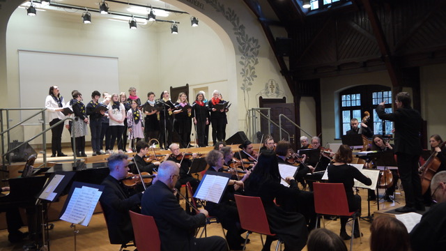 Im Vordergrund sitzen die Musizierenden und spielen auf ihren Instrumenten, im Hintergrund auf der Bühne singt der Chor.
