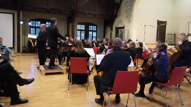 Die Musizierenden sitzen mit ihren Instrumenten in Reihen, die Blicke sind auf den Dirigenten gerichtet.