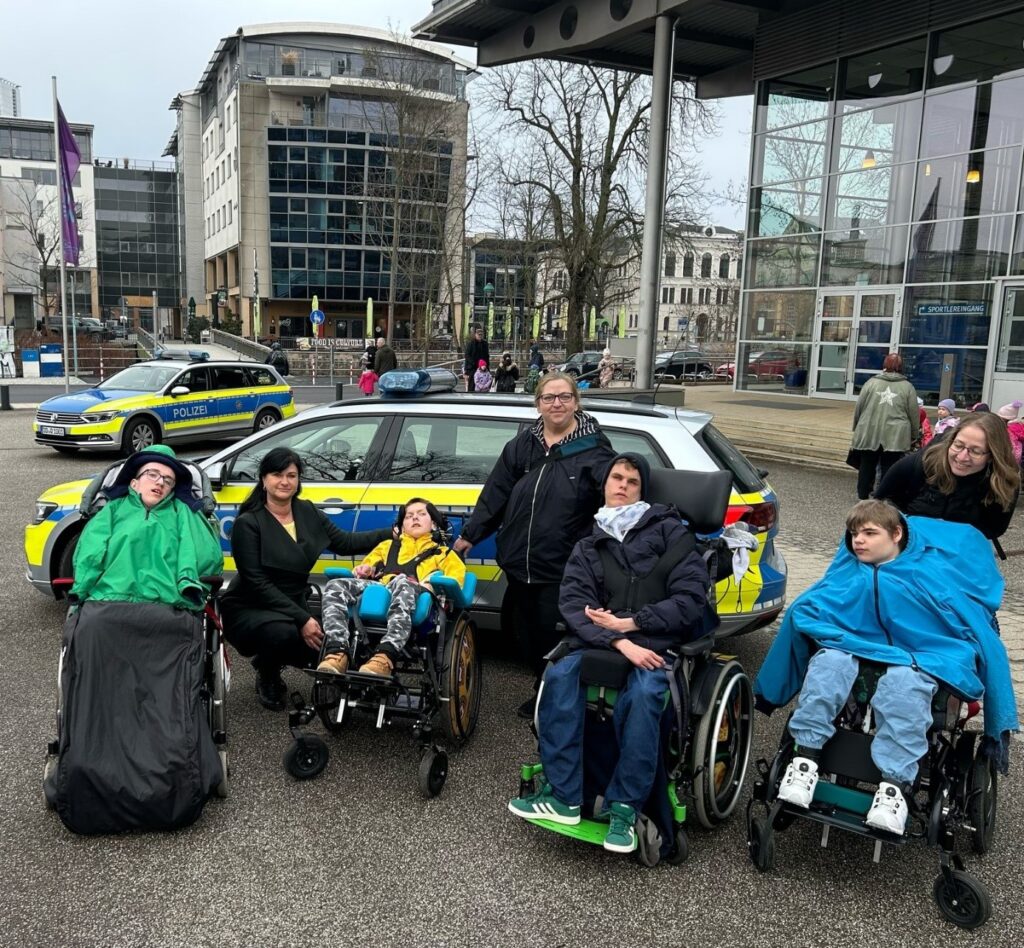Kinder im Rollstuhl und Betreuer vor einemgelb-blau-silbernen Polizeiauto