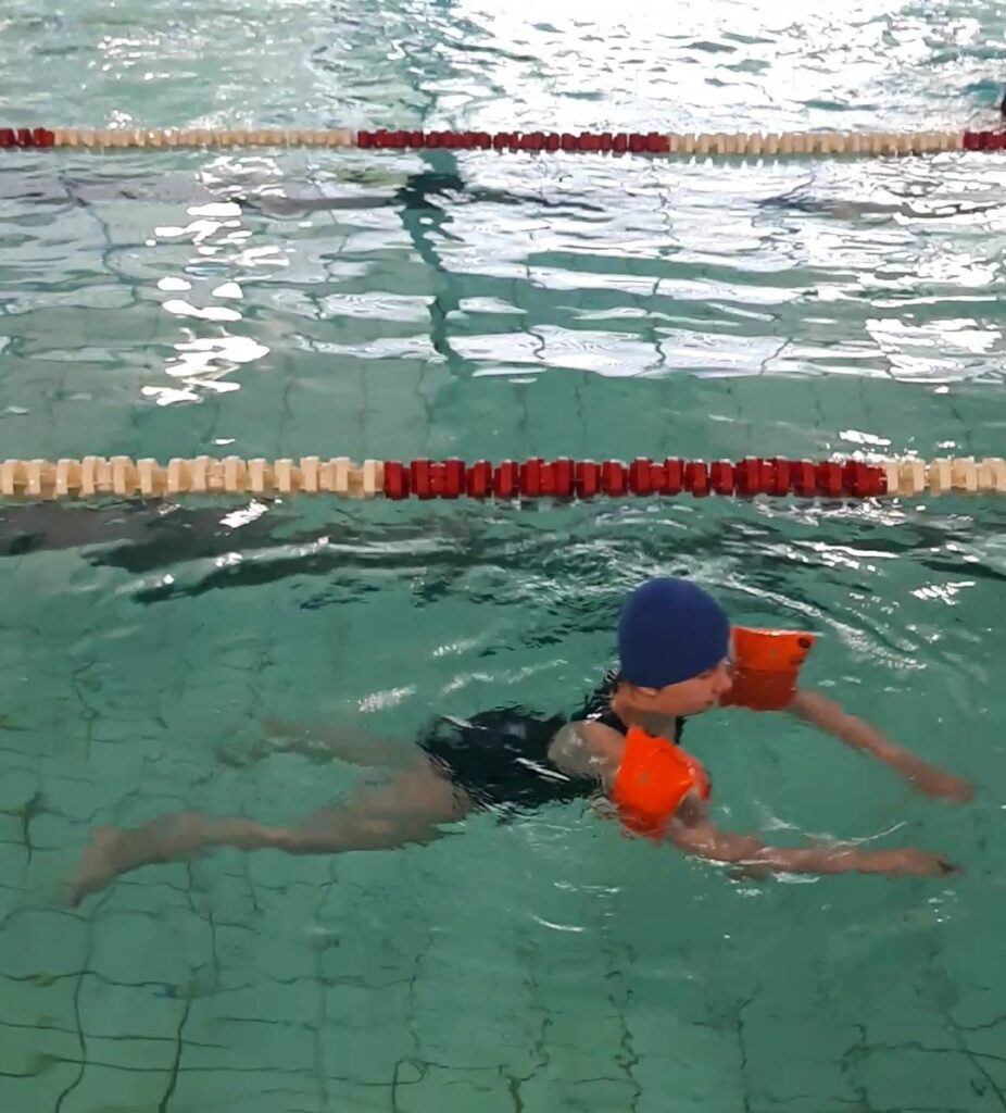 ein Schülerin im Wasser hat zwei Schwimmflügel und eine blaue Badekappe an