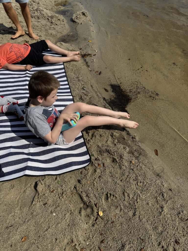 Zwei Schüler liegen am Strand nah am Wasser auf einer Decke, ihre Füße berühren den Sand.