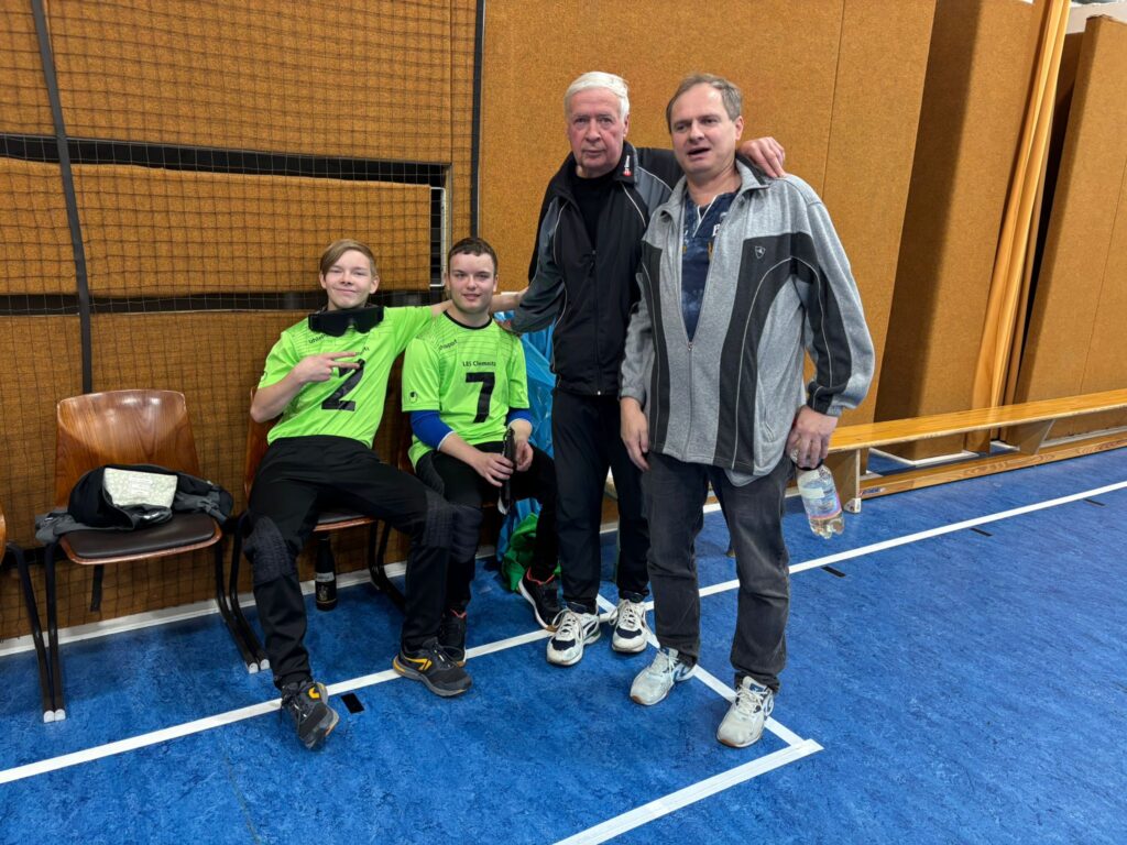 Man sieht zwei Schüler in Trikots, die auf Stühlen sitzen. Daneben stehen die Trainer. Sie sind in einer Turnhalle