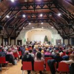Ein großer Saal mit vielen Menschen, die auf Stühlen sitzen und einem Weihnachtskonzert auf der Bühne zuhören