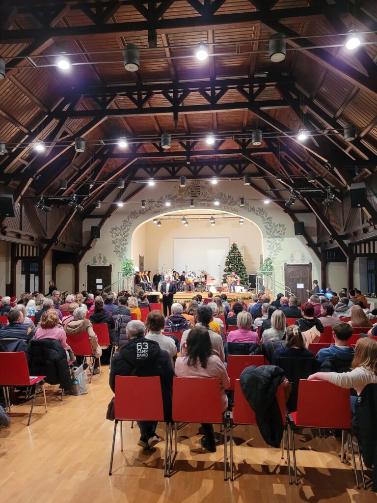 Ein großer Saal mit vielen Menschen, die auf Stühlen sitzen und einem Weihnachtskonzert auf der Bühne zuhören