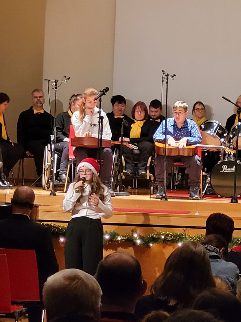 Ein Mädchen singt vor der Bühne mit Mikrofon und dahinter sitzen eine Musikgruppe auf der Bühne.