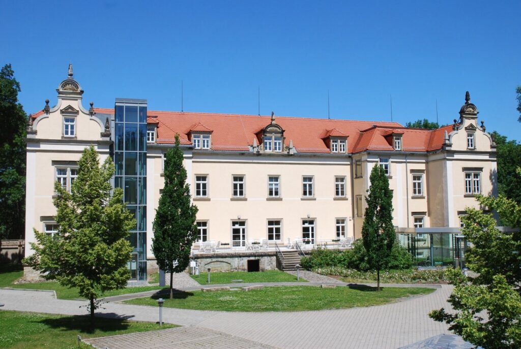 Blick auf das Gebäude der ehemaligen Heil- und Pflegeanstalt Pirna-Sonnenstein. Ein dreistöckiges, helles Gebäude mit rotem Dach. Im Vordergrund Wiese und mehrere Bäume