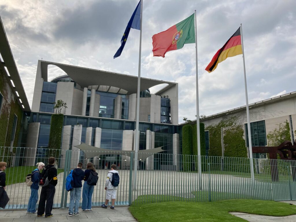 Die Reisegruppe vor dem Bundeskanzleramt. Es wehen drei Fahnen: Deutschland, Europa, Portugal. Bedeckter Himmel