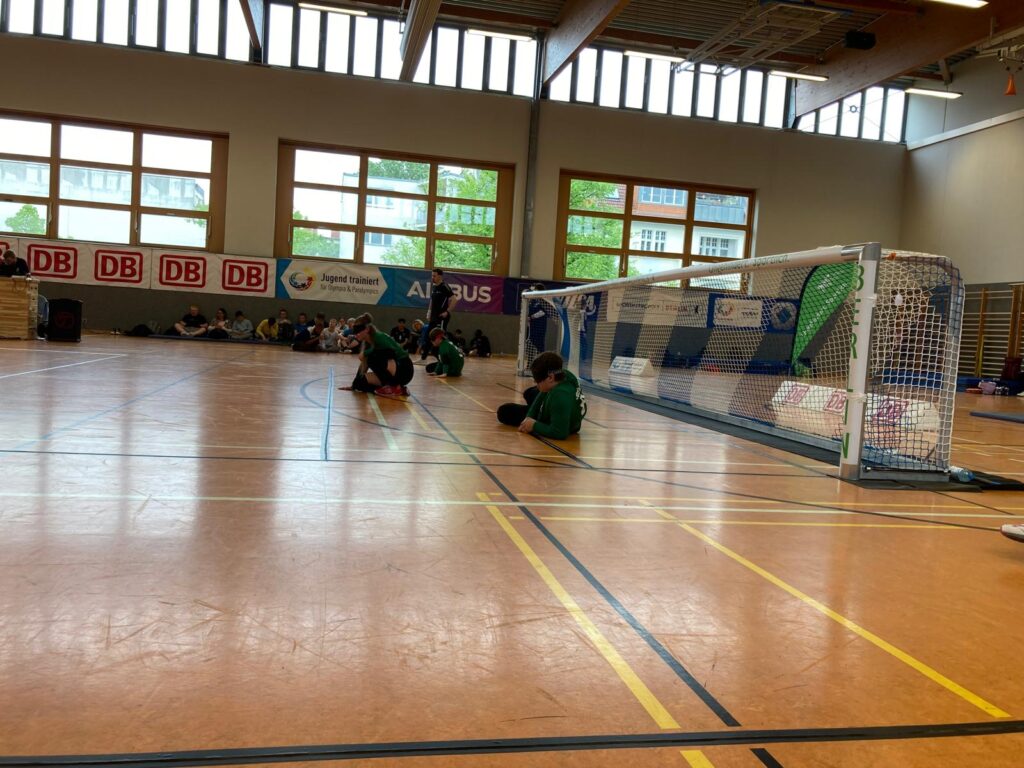 Beim Goalballspiel: Das Team vor dem eigenen Tor in Abwehrposition, alle tragen eine Dunkelbrille. Im Hintergrund sitzen andere Mannschaften am Hallenrand und schauen zu.