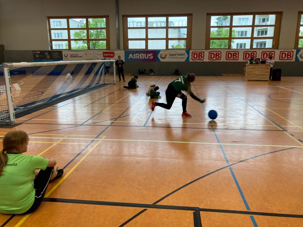 Beim Goalballspiel: Die Schülerin im Vordergrund spielt gerade den Ball, im Hintergrund ihre Teammitglieder in Abwehrposition. Vorne links im Anschnitt die Trainerin auf dem Boden sitzend.