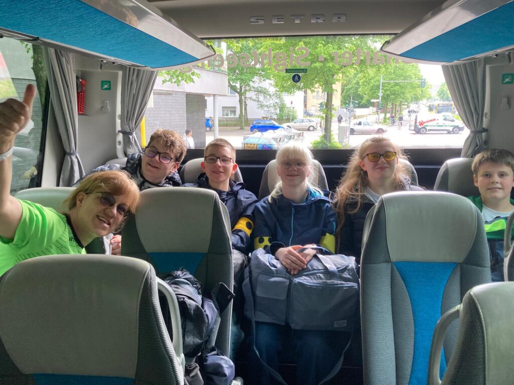 5 Schülerinnen und Schüler und eine Lehrerin sitzen in einem Bus. Die Lehrerin zeigt den Daumen nach oben, alle lachen.