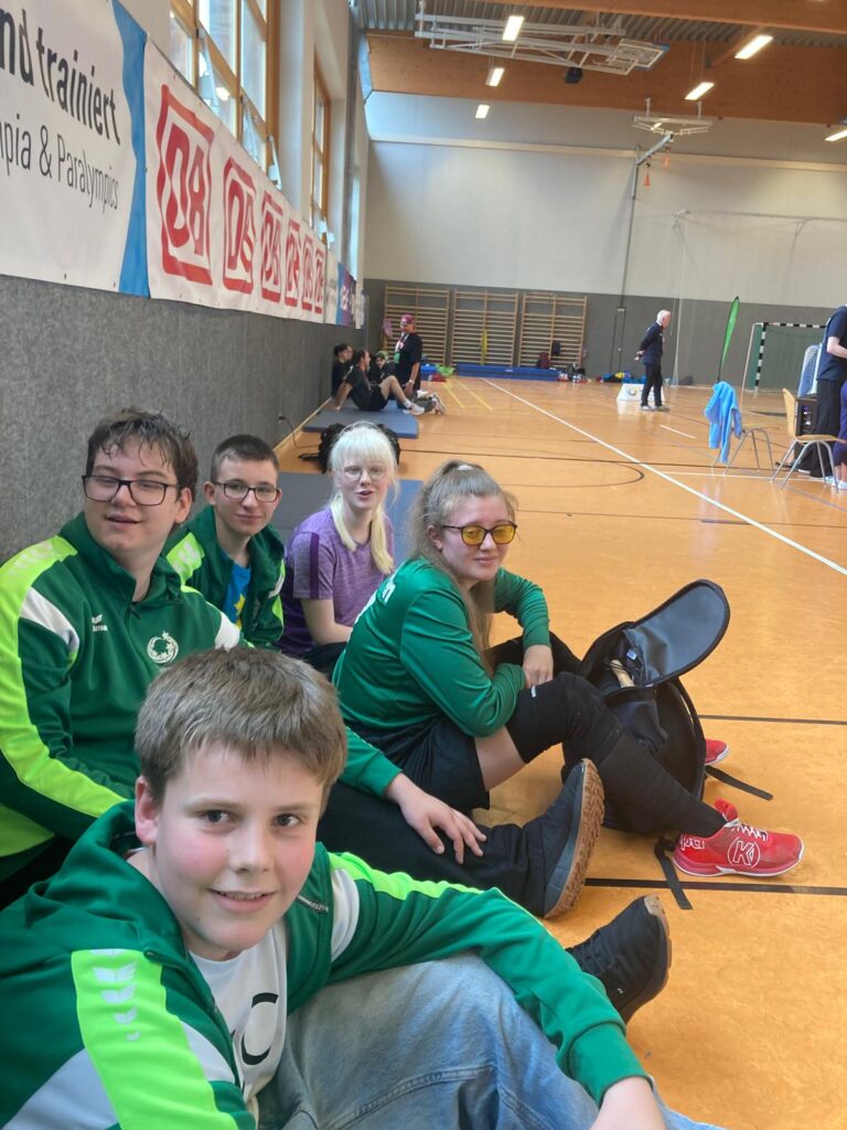 Die Schulmannschaft aus 2 Schülerinnen und 3 Schülern sitzt in der Sporthalle am Rand auf dem Boden. Im Hintergrund Mitglieder einer anderen Mannschaft.
