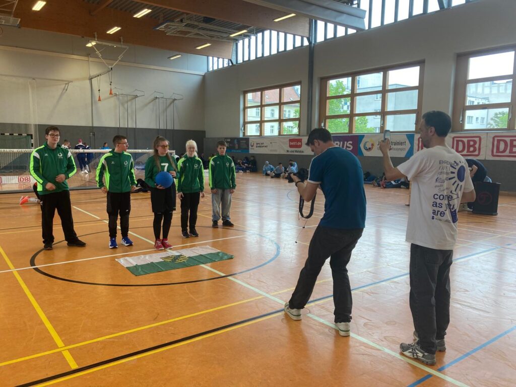 Das Team wird in der Turnhalle auf dem Spielfeld von zwei Personen gleichzeitig fotografiert. Eine Person fotografiert mit einem Fotoapparat, eine mit einem Smartphone. Die Spielerin in der Mitte hält einen blauen Ball in den Händen.