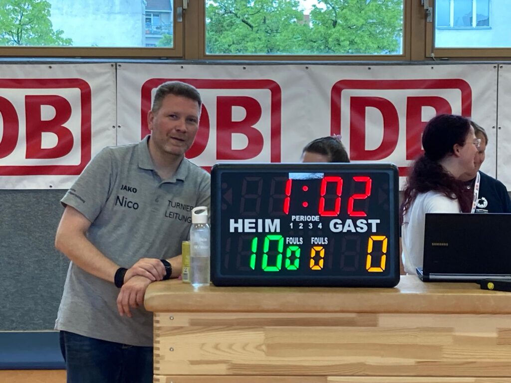 Ein Mann steht neben einer Anzeigetafel. Die Restzeit beträgt 1:02 Minuten. Es steht 10:0 für das Heimteam.