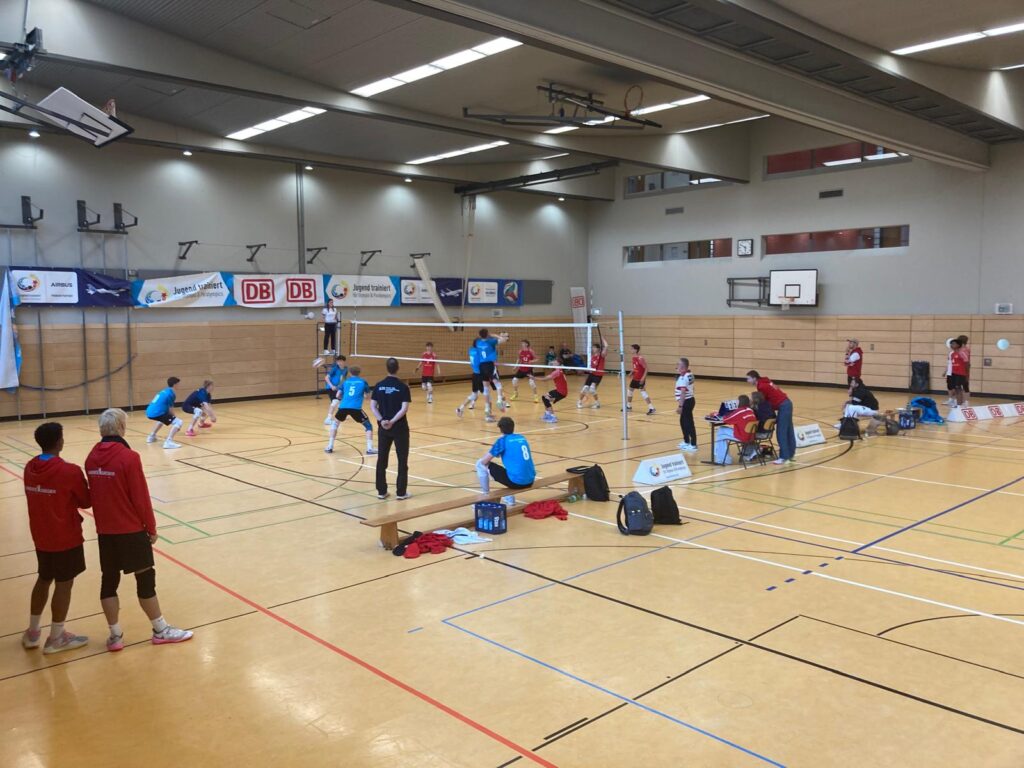 In einer Turnhalle spielt ein blaues Team Volleyball gegen ein rotes Team.