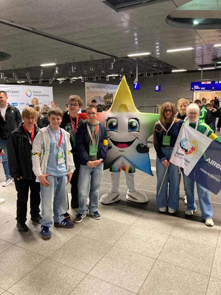 Gruppenfoto der Reisegruppe aus 5 Schülerinnen und Schülern sowie 2 Lehrerinnen in einer Bahnhofshalle. Zwischen ihnen eine Person in einem Kostüm, dessen Oberteil die Form eines Sterns mit Gesicht und bunten Zacken hat. Die Schülerinnen halten eine Fahne "Jugend trainiert" in der Hand.