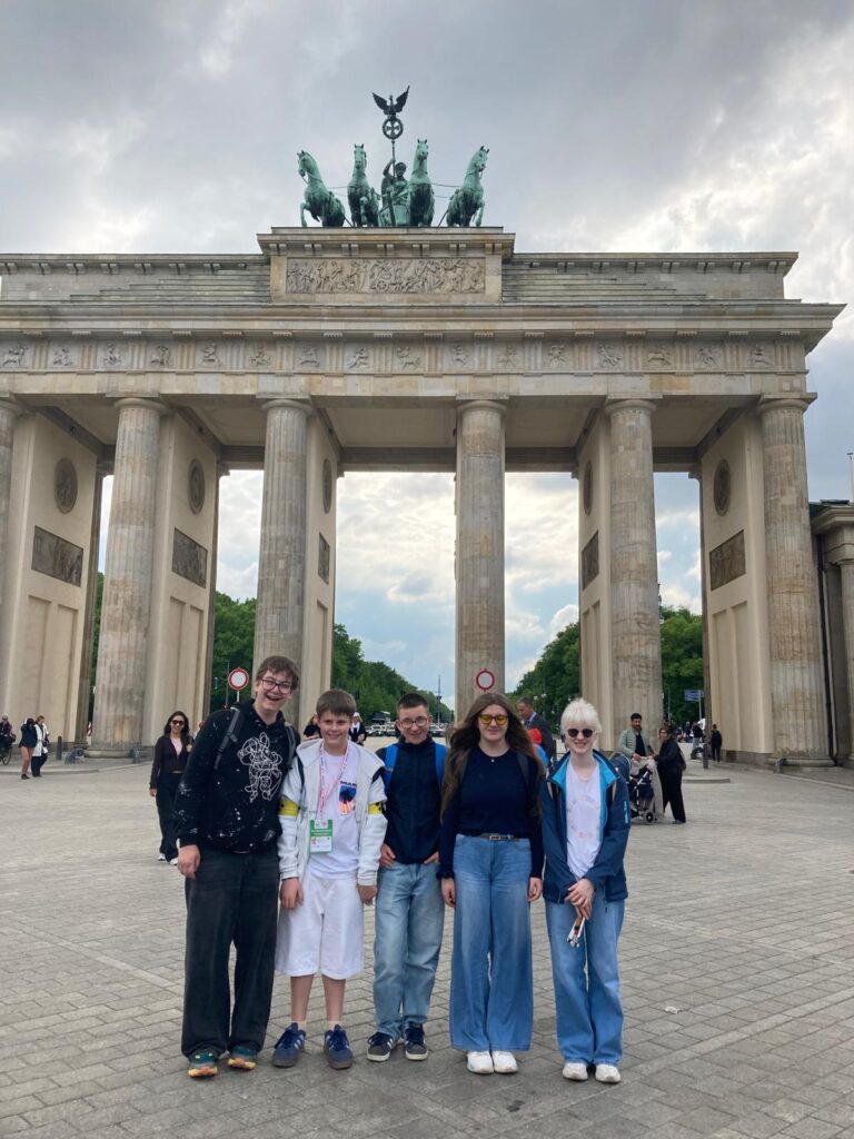Gruppenfoto der fünf Schülerinnen und Schüler vor dem Brandenburger Tor, bedeckter Himmel. Alle lächeln.