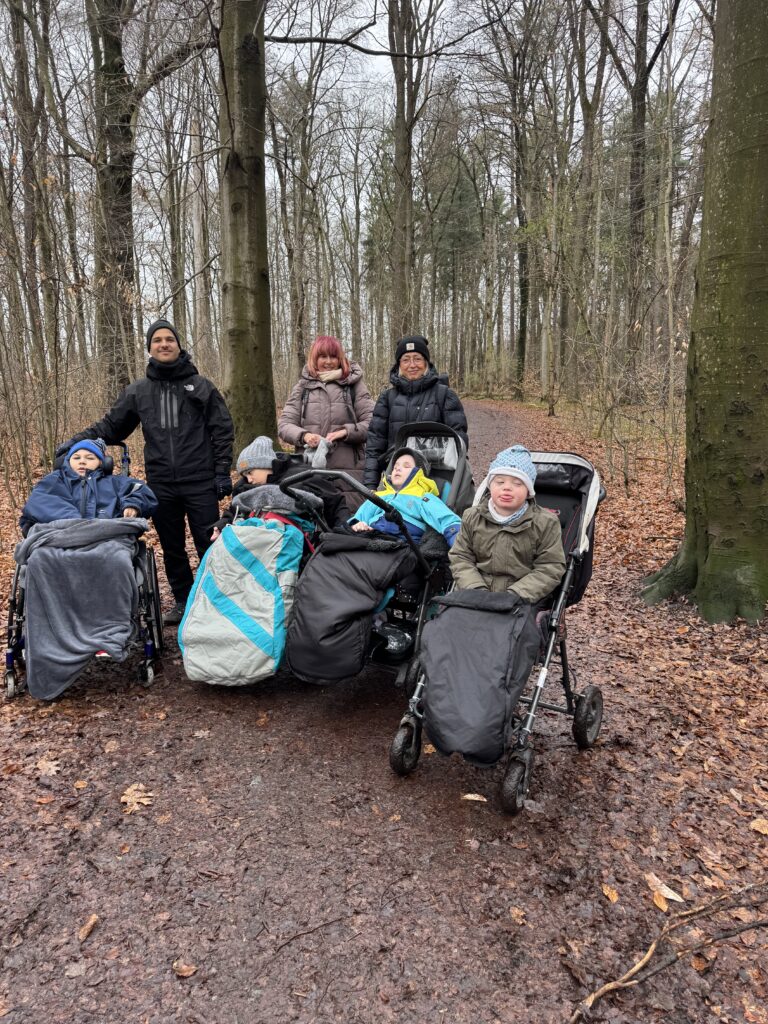 Gruppenfoto im Wald. Die Schüler sitzen in ihren Buggys bzw. Rollstühlen, dahinter steht das Klassenteam (ein Mann, zwei Frauen). Alle tragen eine Jacke, viele haben eine Mütze auf.