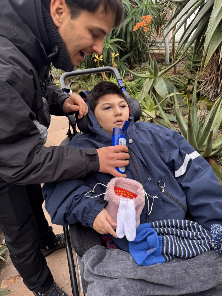 Ein Schüler im Rollstuhl. Ein Lehrer beugt sich über den Schüler und hält ihm eine offene Tube unter die Nase. Der Schüler hält ein gefundenes Osternest in der Hand.