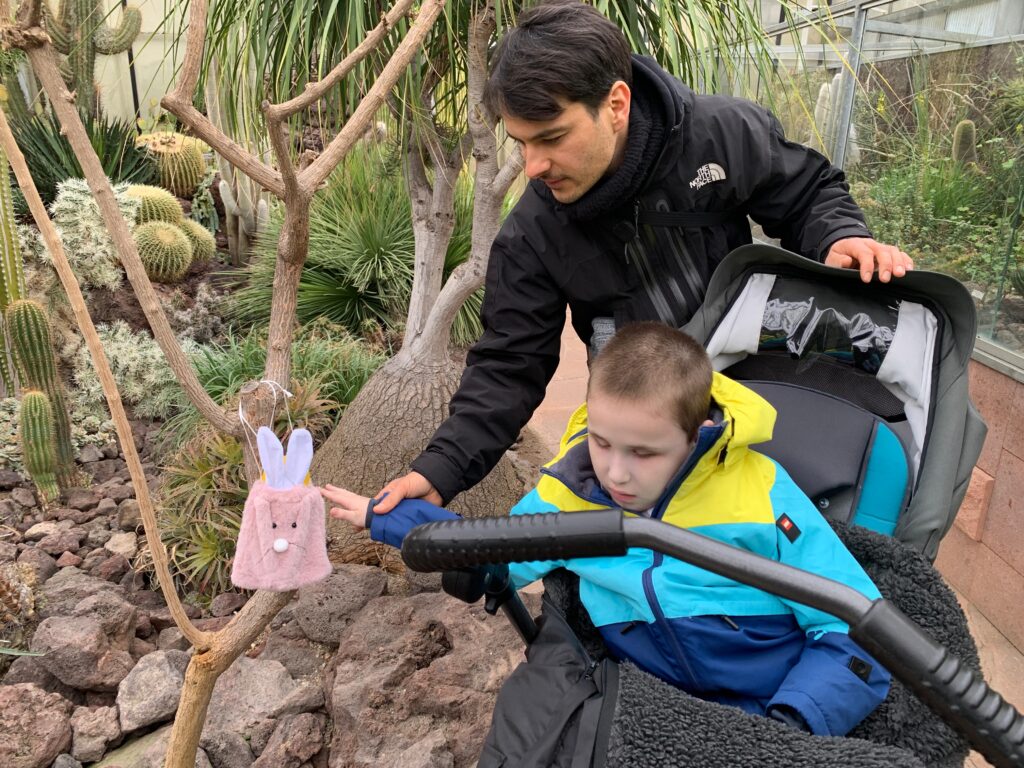 Ein Schüler im Buggy im Innenraum des Botanischen Gartens. Ein Lehrer steht hinter ihm und führt seine Hand zu einem im Baum hängenden Osternest.