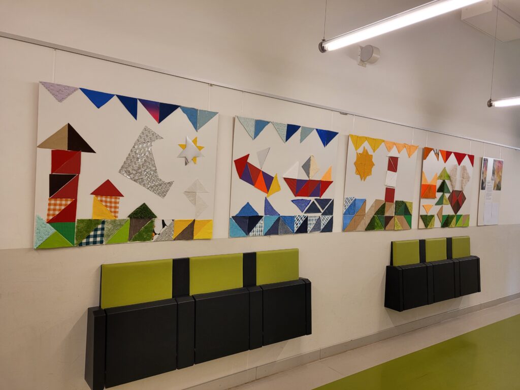 Ein zusammenhängendes Bild, welches aus vier einzelnen Teilen besteht, hängt an einer Wand auf dem Schulflur. Darunter befinden sich zwei mal drei Klappsitze an der Wand.