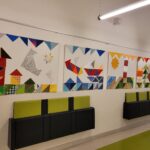 Ein zusammenhängendes Bild, welches aus vier einzelnen Teilen besteht, hängt an einer Wand auf dem Schulflur. Darunter befinden sich zwei mal drei Klappsitze an der Wand.