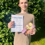 der Schüler Ricardo hält eine Urkunde in der Hand mit dem 1. Platz und einem Gutschein
